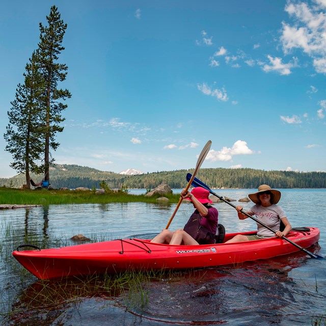 Two women sit in a red kayak on a lake backed by a snow-capped mountain