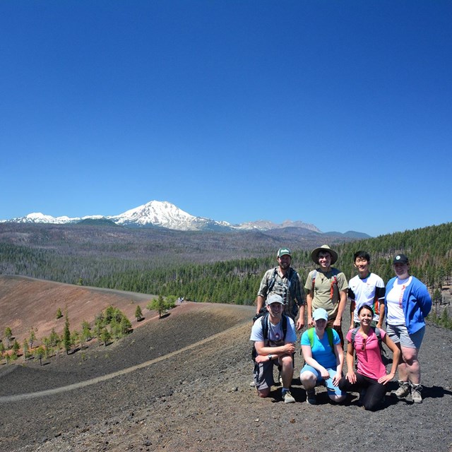 A group of hikers poses for a photo on the summit of a black ridge backed by a snow-capped volcano