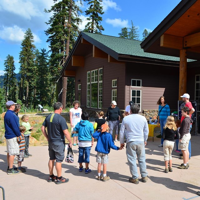 A group of people stand in a circle while conducting an activity outside of a wood building