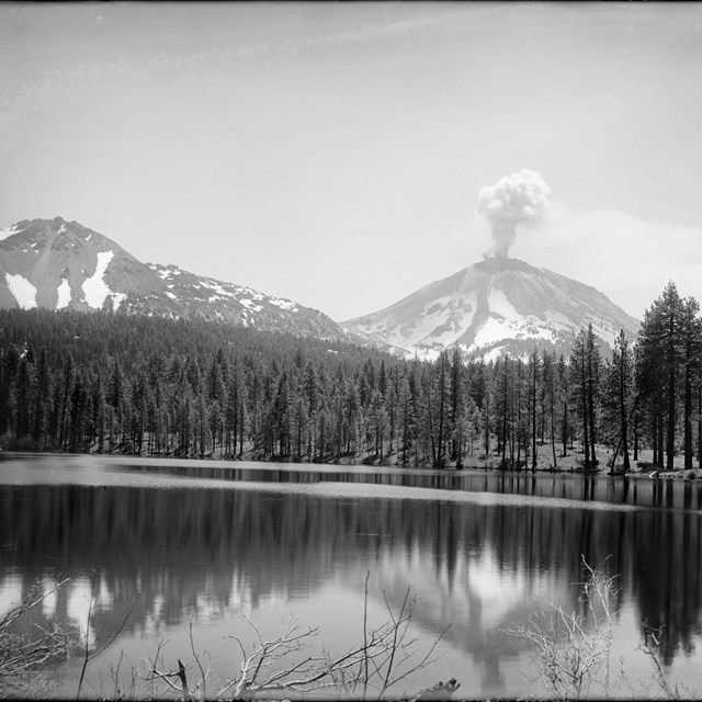 Black and white photo of a volcano with a small ash cloud fronted by a lake