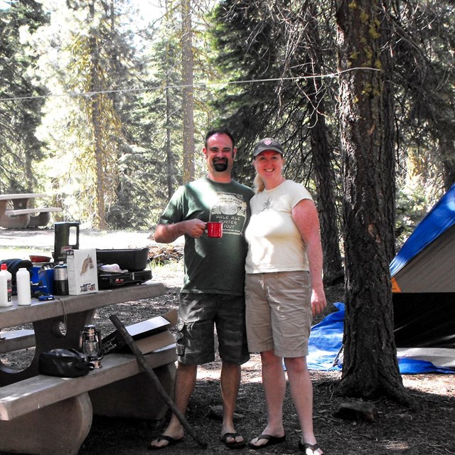 A man and a woman standing next to a picnic table and tent at a campground.