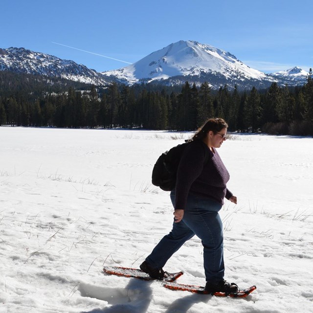 Two women on snowshoes walk across the shore of a snow-covered lake backed by volcanic peaks.