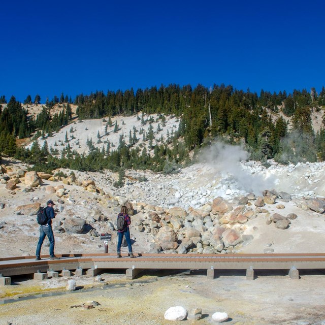 A man and a woman walk along a boardwalk in a hydrothermal area. 