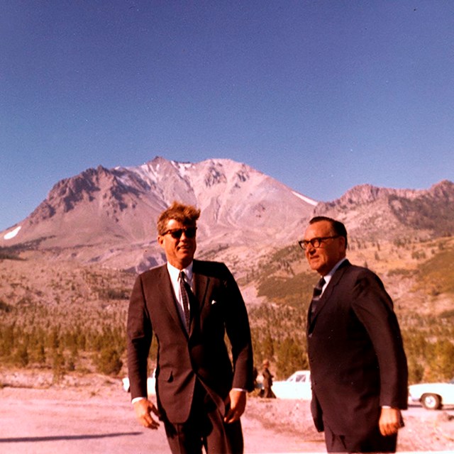 Two men in black suits standing backed by a barren volcanic peak.
