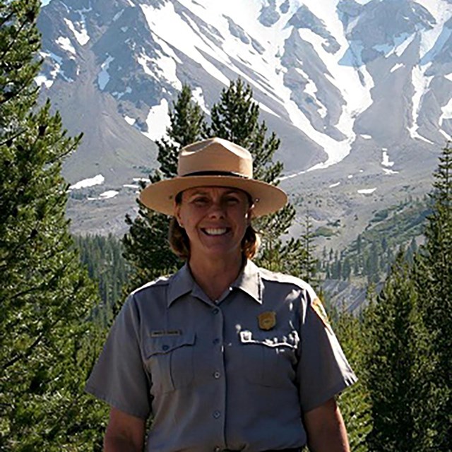 A women in a ranger uniform stands in front of a barren volcanic peak