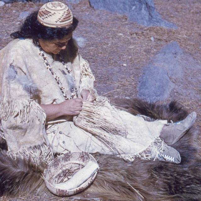 A woman in a leather shirt and skirt weaves a basket while sitting on animal furs.