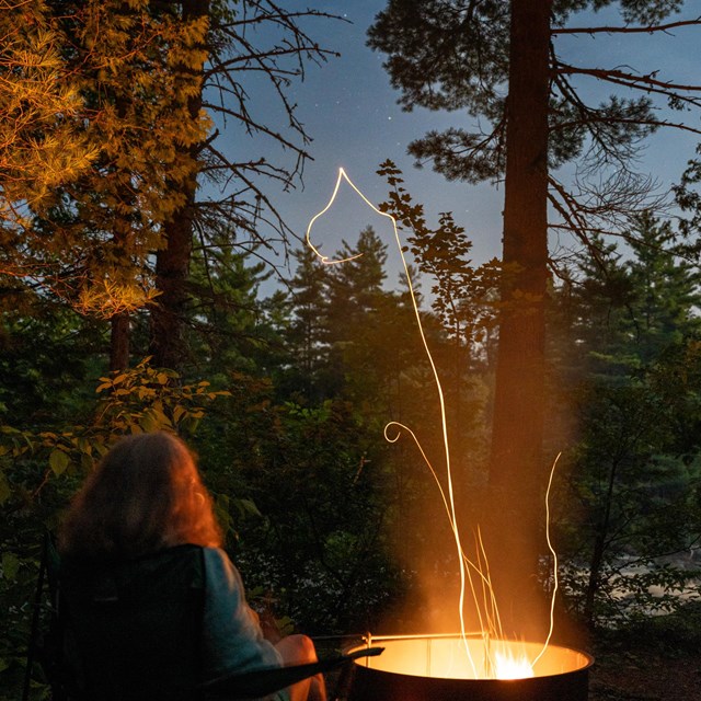 a fire in a metal fire ring at night, a woman sitting next to the fire, stars and trees overhead