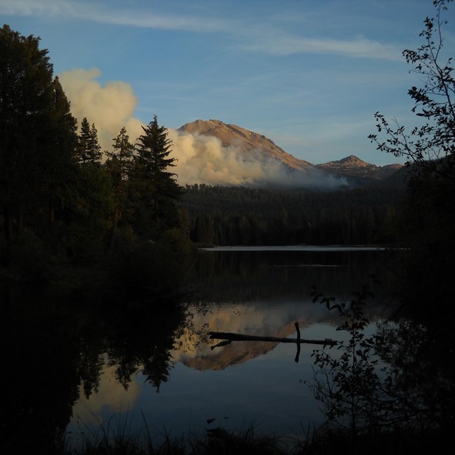 smoke plumes across a mountain, reflected in the lake below