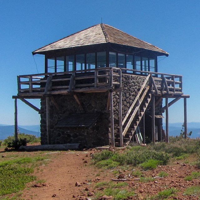 A two-story stone and wood fire lookout on the top of a rocky hill backed by distant mountains.