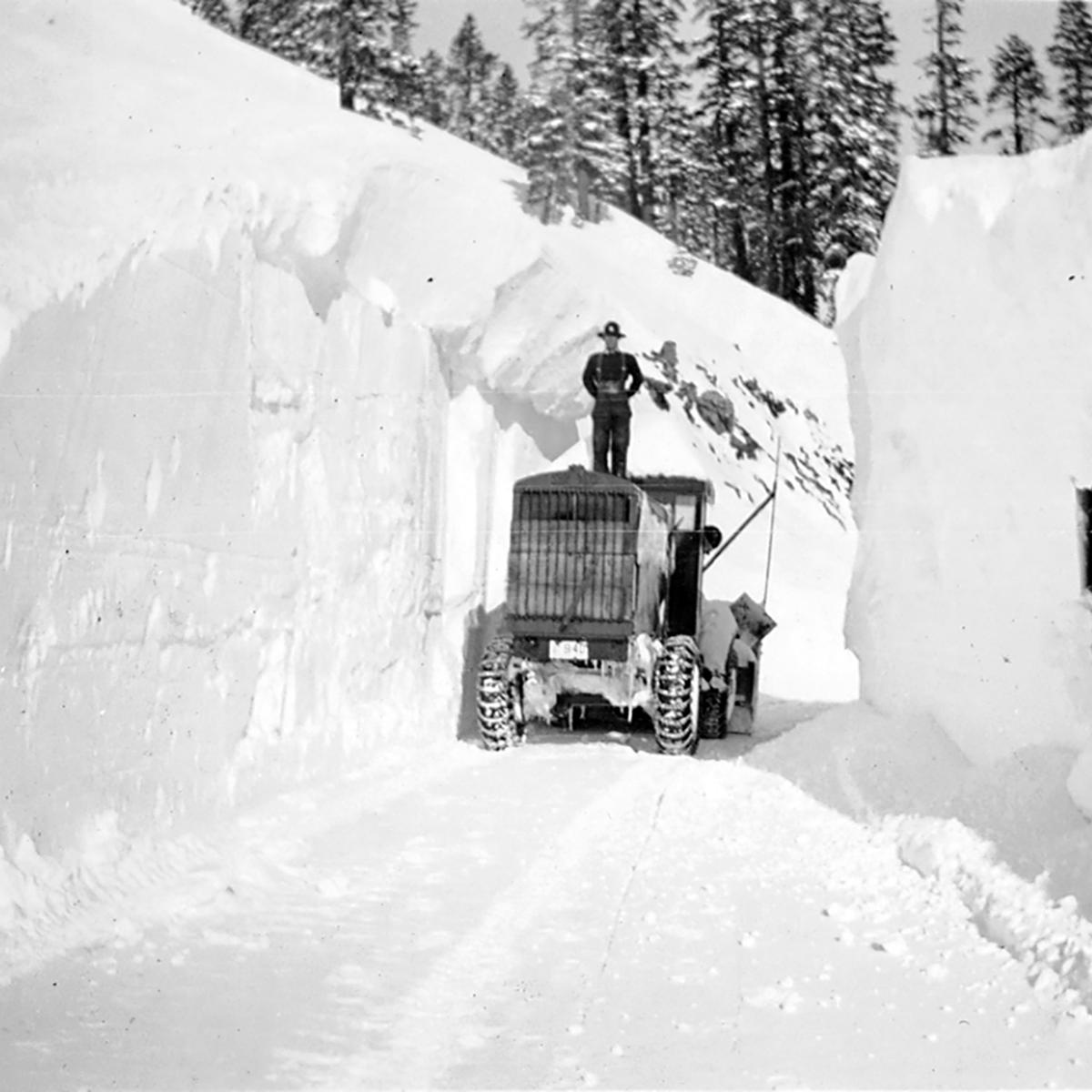 A man stands on a snowplow flanked by walls of snow