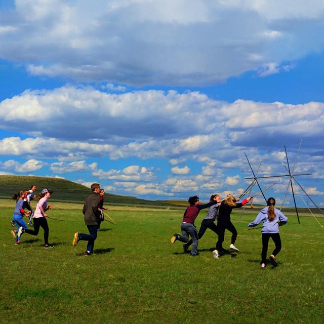 Group of teenagers run to the right playing a game. Blue sky. Green field. Teepee frame behind.