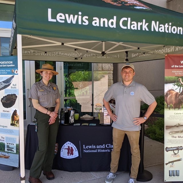 Two rangers stand outside next to tent which reads Lewis and Clark National Historic Trail