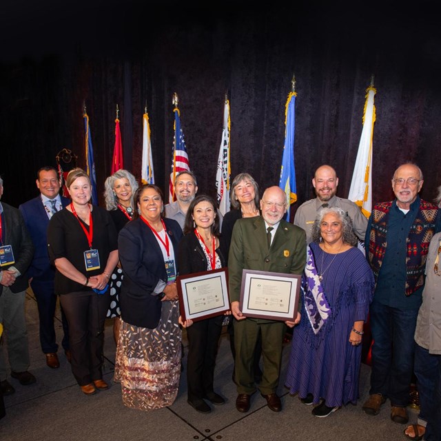 13 people pose around a certificate. Some wear Park Service Uniforms One wears tribal print vest