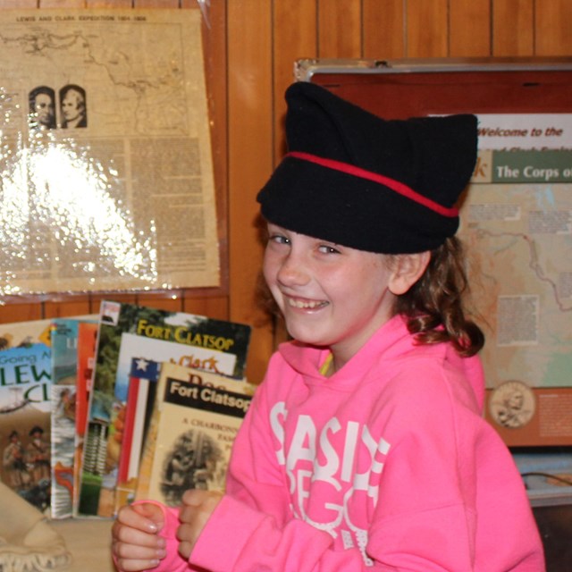 Girl wears a fatigue hat in front of traveling trunk materials. 