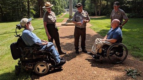Three park rangers with two men in wheelchairs talking outside.