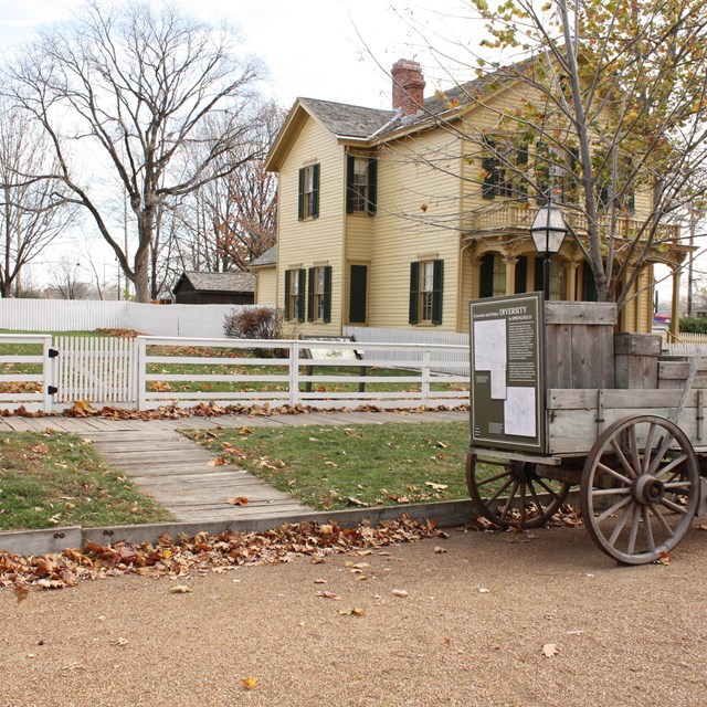 Empty grass lot surrounded by white fence, with recreated wagon on street in front of lot