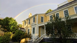 Large yellow Georgian house with green trees in foreground and rainbow overhead.
