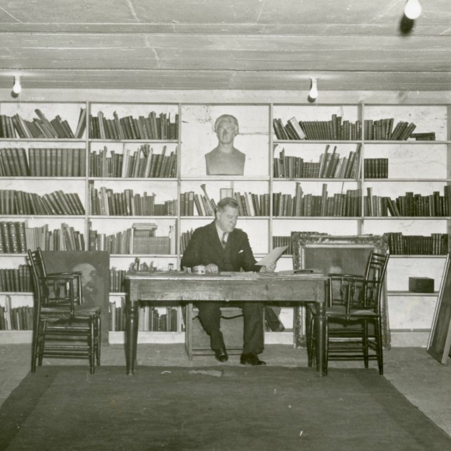 black and white photograph of man seated at desk, full wall of bookshelves behind him with bust