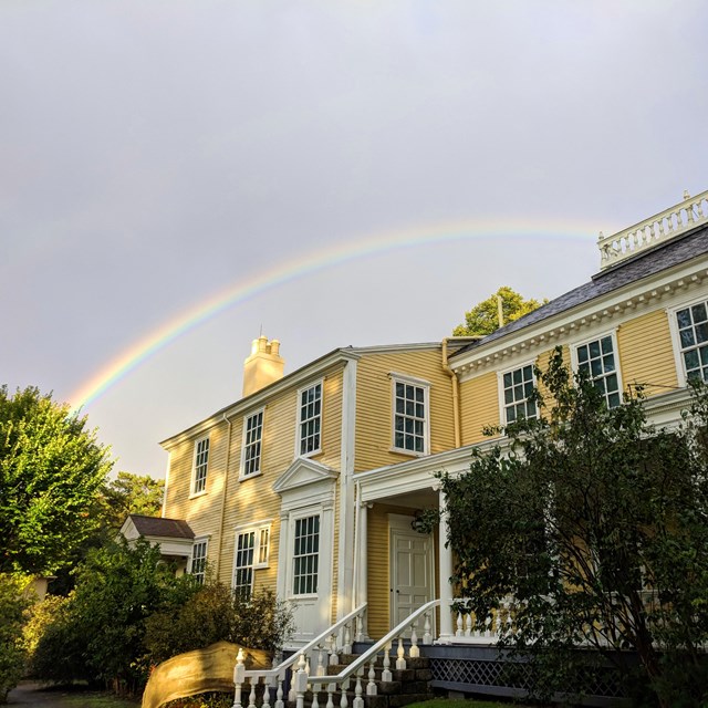 Large yellow Georgian house with green trees in foreground and rainbow overhead.