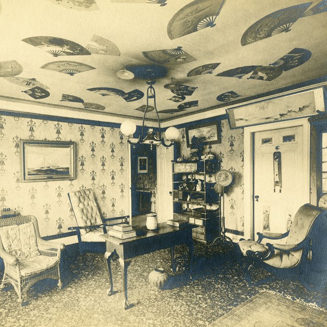 Black and white photograph of room with several chairs and paintings, ceiling covered with fans.