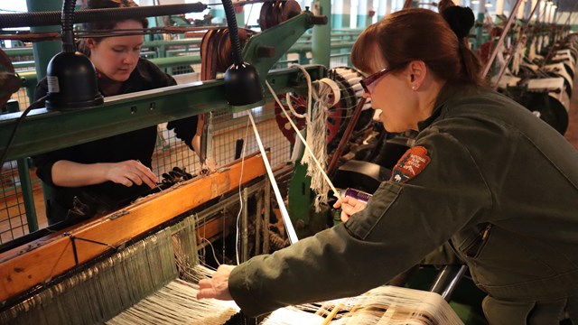 Two employees work on a loom, drawing in the yarn.