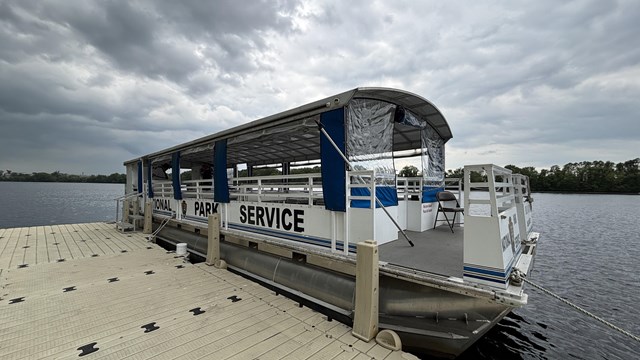 A close top pontoon boat emblazoned with the words "National Park Service" is tied up at a dock