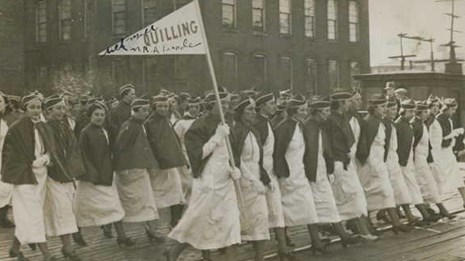 Women dressed in white marching in a parade holding flags