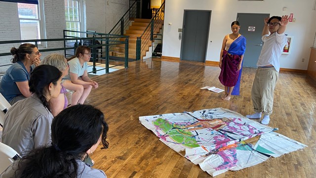 A group of people sit around tables painting sunflowers and smiling.