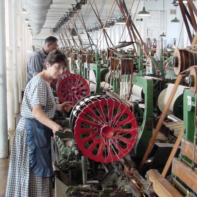 Two workers, a man and a woman, work on a line of power looms in a recreated factory setting