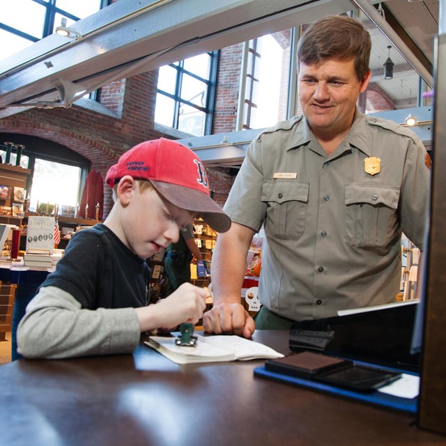 A park ranger helps a young boy stamp his passport book
