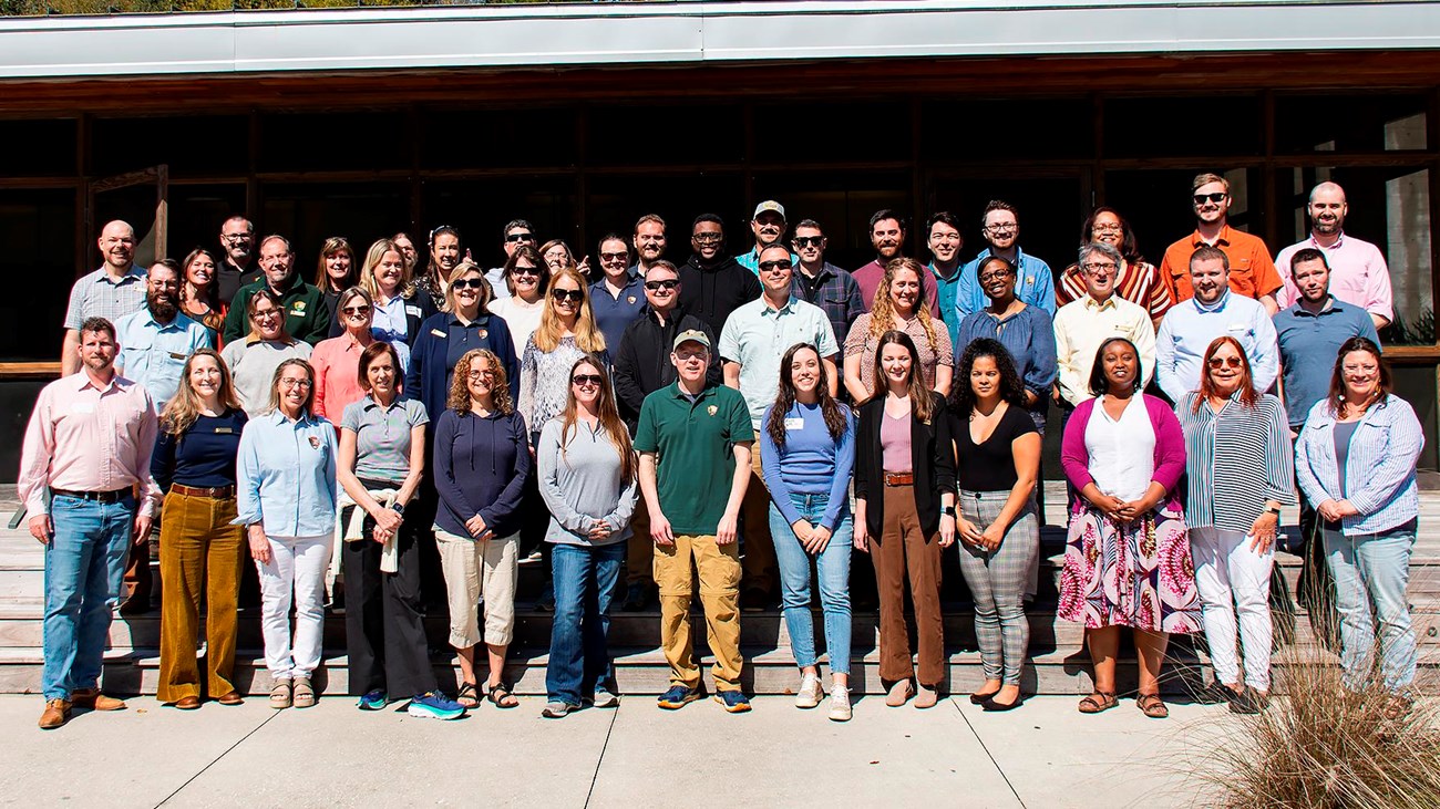 a smiling group of 40 people all wearing different colors stand in a rows for a team photo. 
