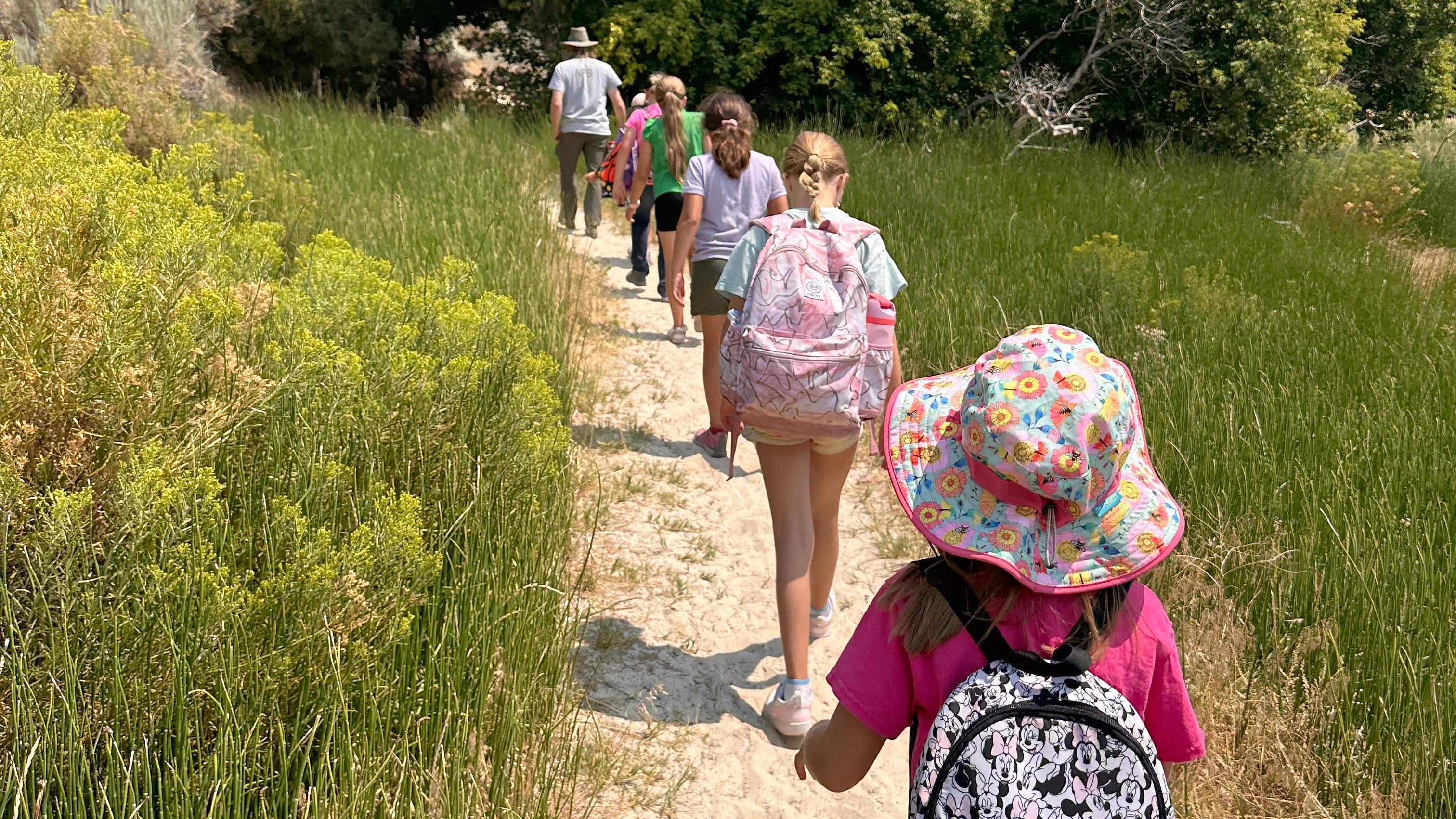 Children with colorful packs being led on a dirt path with green vegetation, trees, blue skies