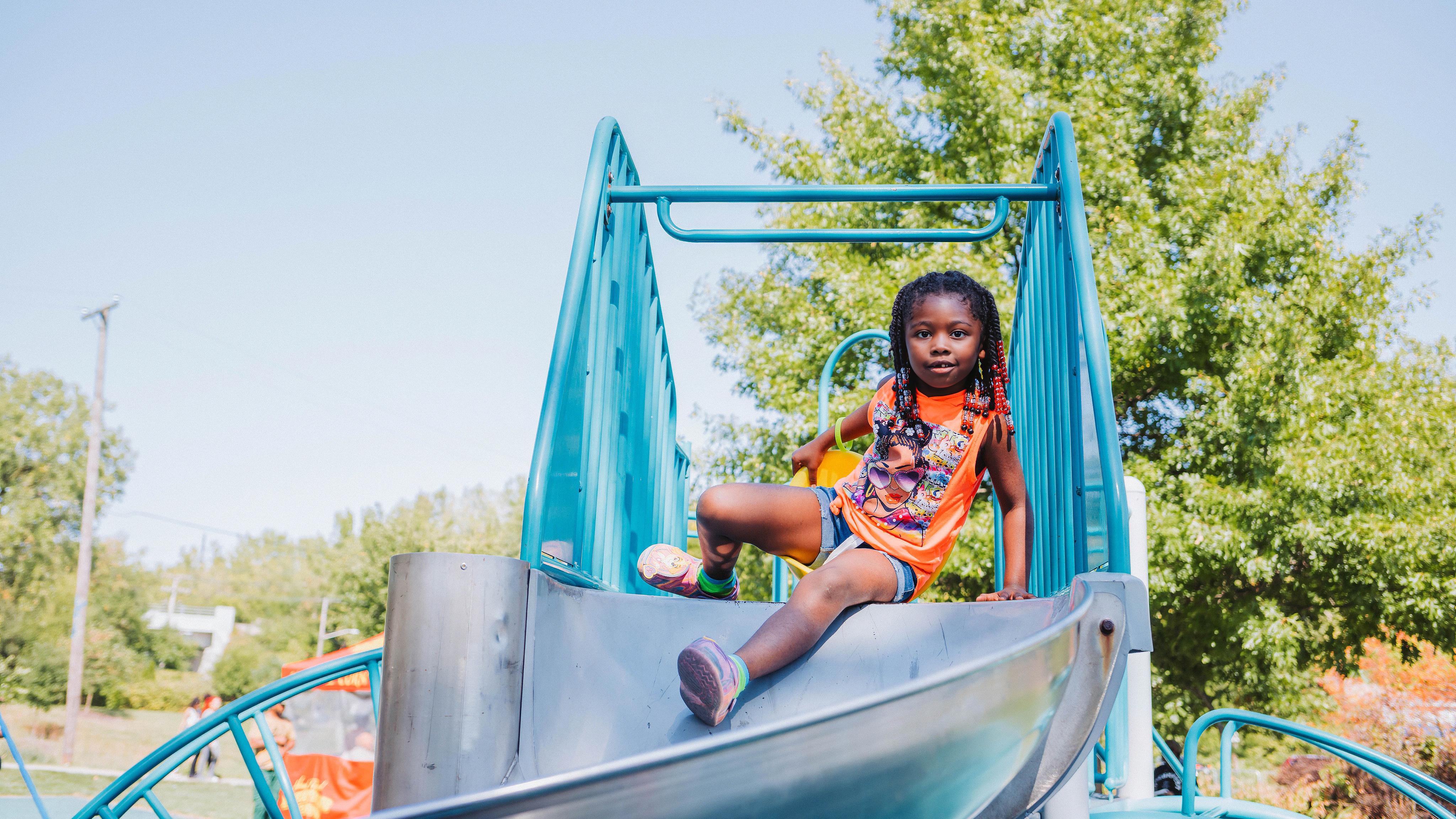 Girl with braids on a colorful playground slide, trees, blue skies in background
