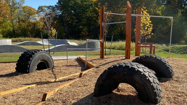 A blue sky above an adventure park with large rubber tires half buried & wooden balance beams.