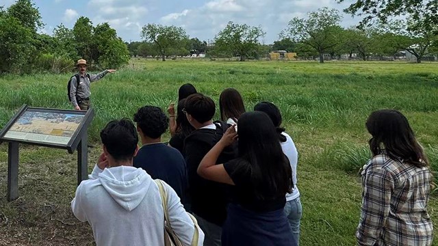A ranger stands in front of a group of people in an open, grassy field under blue skies.