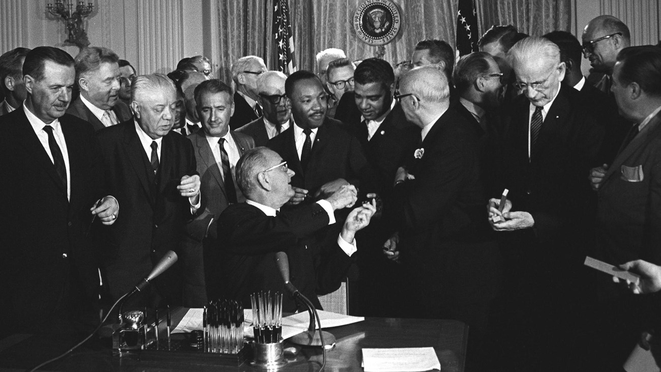 A crowd of men stand behind a desk as President Johnson, seated, hands pens to civil rights figures.