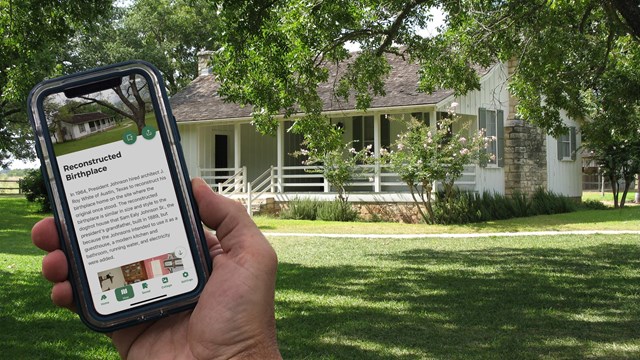 A hand holds a cell phone showing a screen "Reconstructed Birthplace." The home is seen behind.