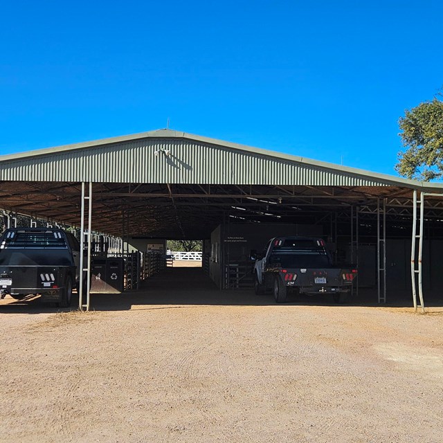 Green barn with ranch vehicles parked in front. Walk way in middle of barn.