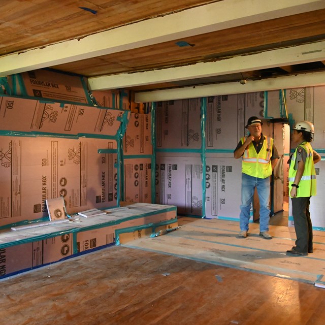 Two people in bright yellow vests stand in a room of cardboard-protected walls.