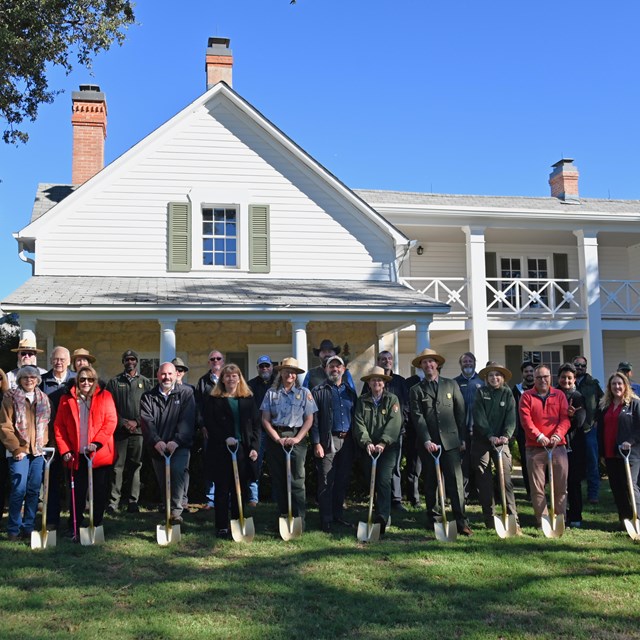 A large group of people, some holding shovels, stand in front of a two-story, white frame house.