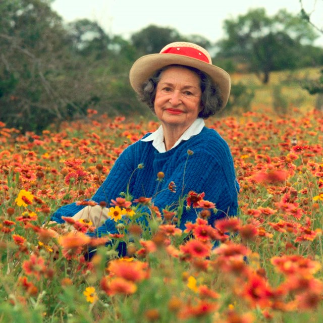An older lady wearing a straw hat sits in a field of yellow and orange flowers.