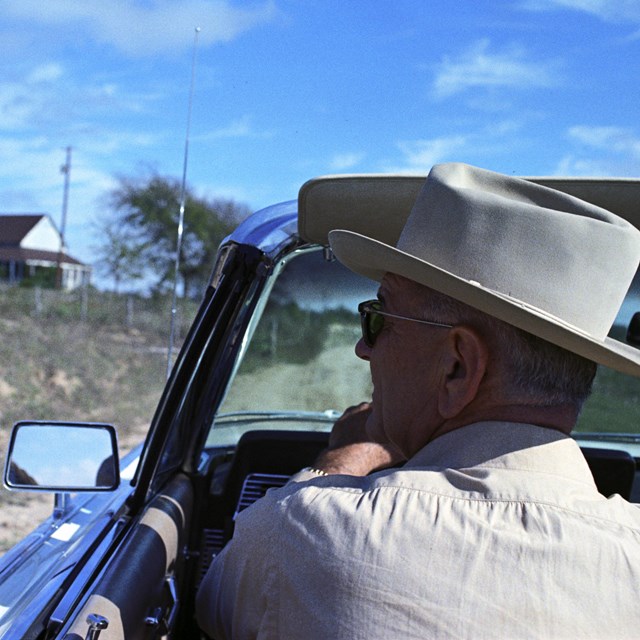 A view from the back seat of a convertible with Lyndon Johnson in the front driver's seat.