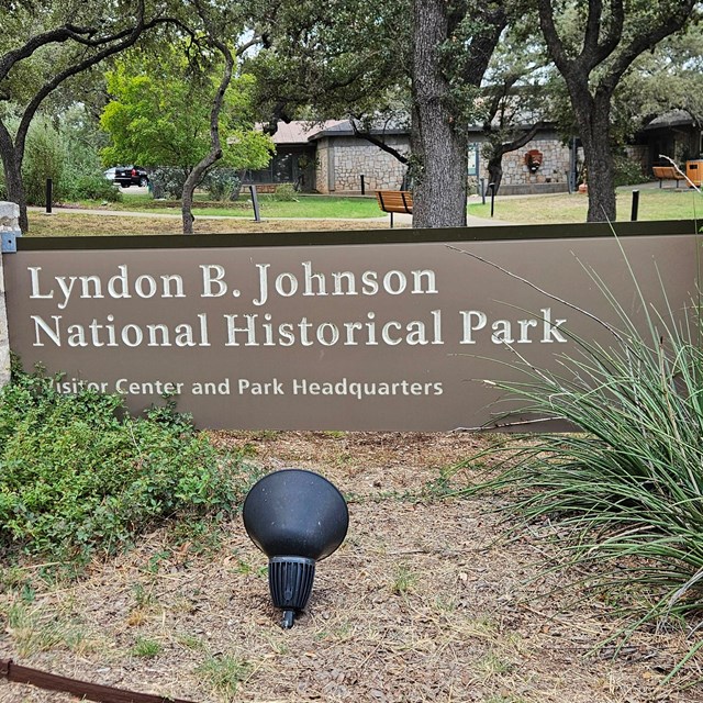 Park sign with bushes on either side. Visitor center building in background.