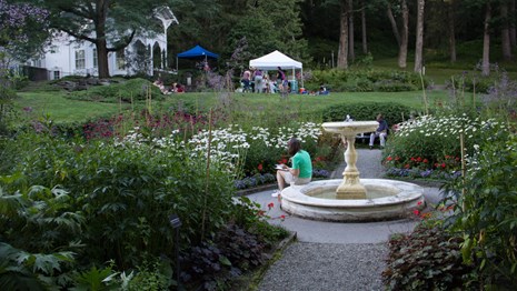woman sits on fountain and paints in formal four square garden, with tents and people in distance