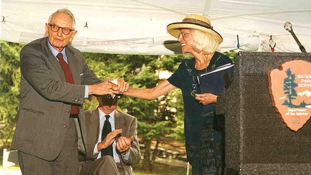 A man in a suit in his 90s shakes hands with a woman standing at a National Park Service podium