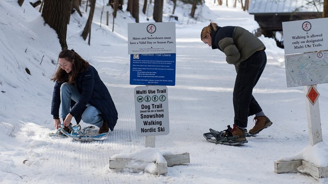 Two people put on snowshoes in front of trail signs and groomed trail