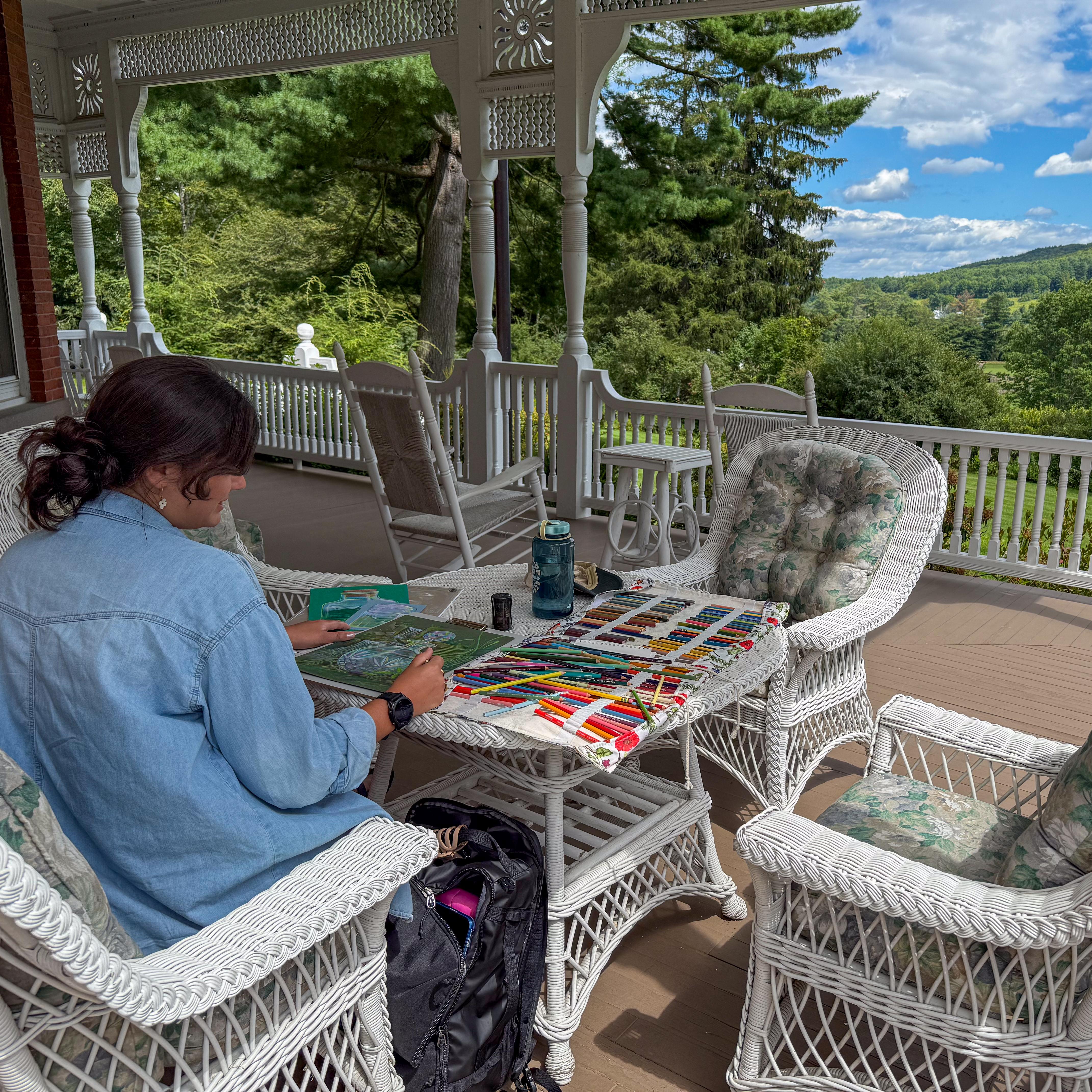 artist sits on MBR mansion porch on wicker furniture with colored pencils on table