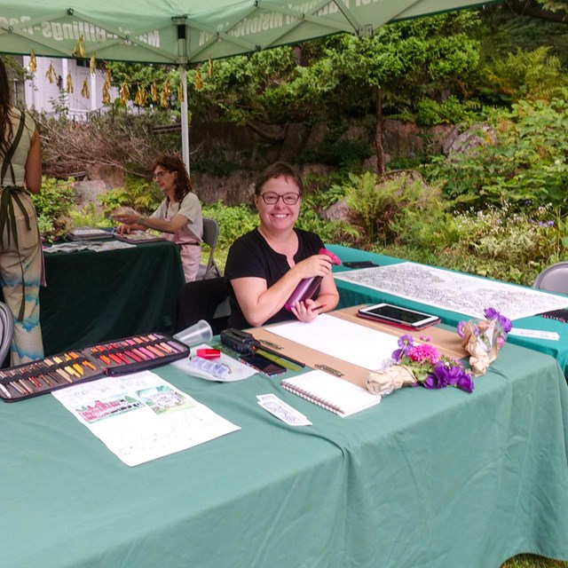 photo of person sitting at an event booth and smiling at camera surrounded by art materials