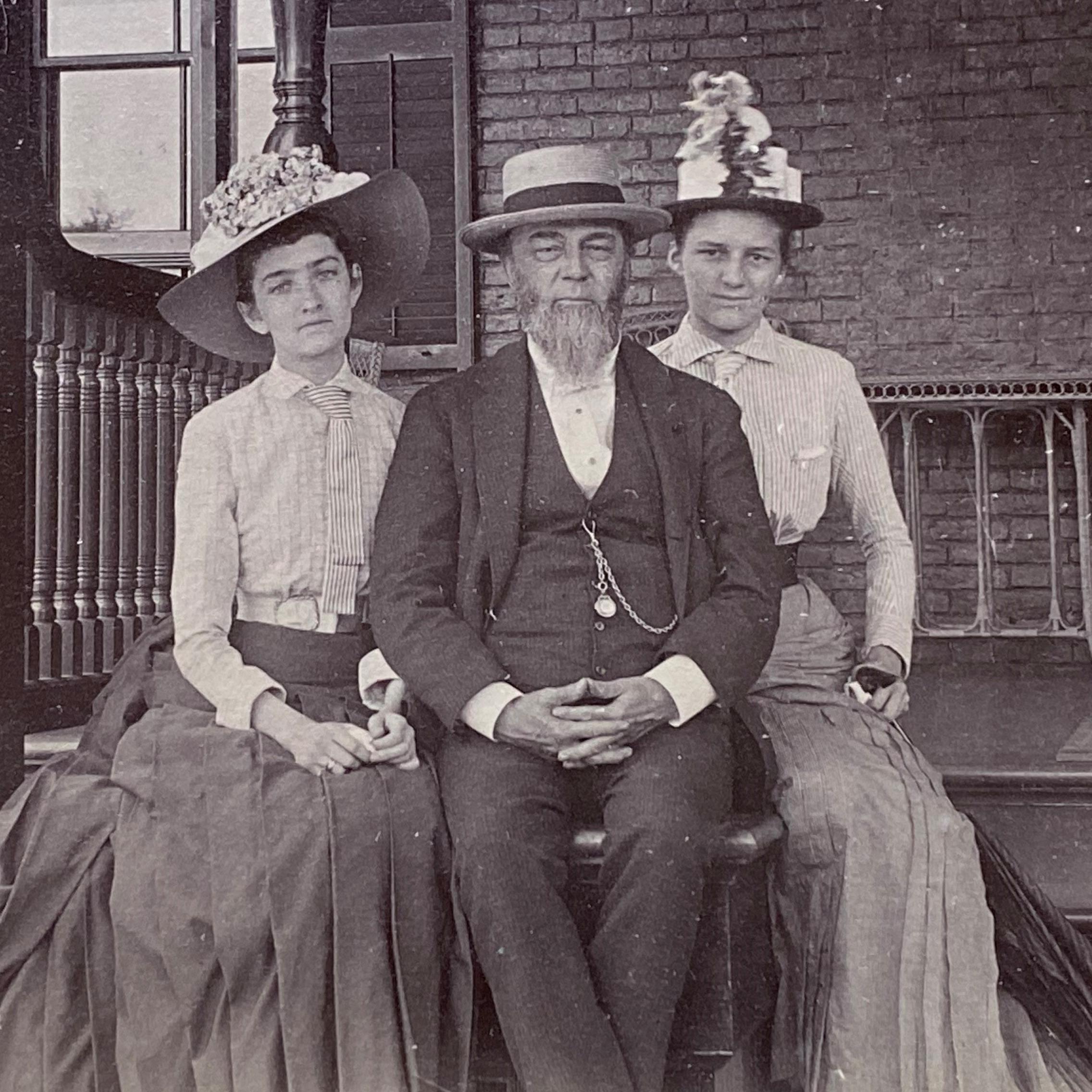 historic black and white photo of older man on sitting between two younger women on porch