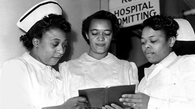 Black and white photo of three women in nurses uniforms looking at a book. 